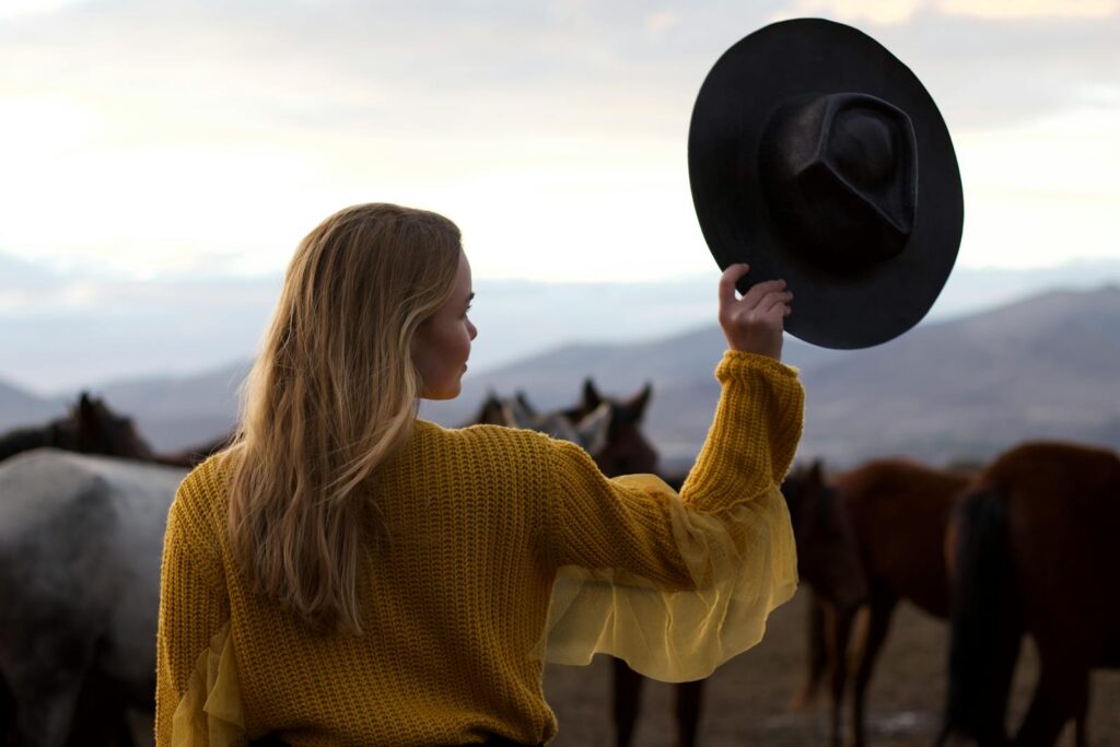 Blonde woman in a knitted sweater holding a black hat, standing in a countryside field with horses.