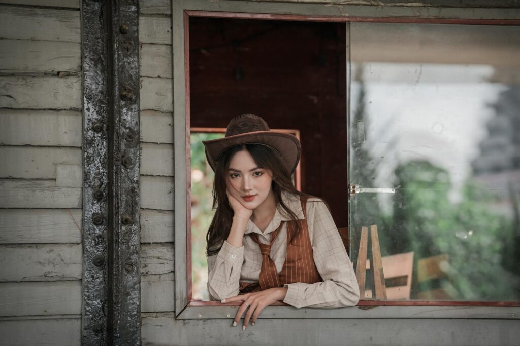 A woman in a vintage western outfit poses thoughtfully by a rustic window.