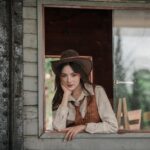 A woman in a vintage western outfit poses thoughtfully by a rustic window.