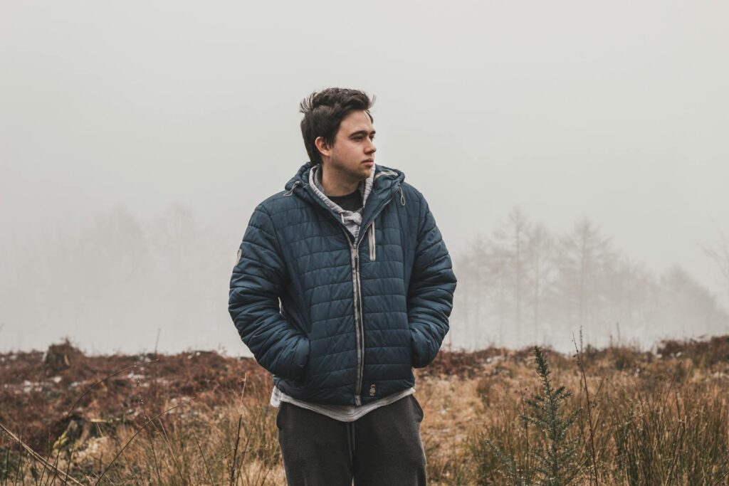 A young man stands in a foggy outdoor setting, wearing a blue jacket and looking contemplative.