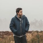A young man stands in a foggy outdoor setting, wearing a blue jacket and looking contemplative.