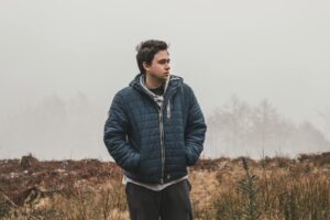 A young man stands in a foggy outdoor setting, wearing a blue jacket and looking contemplative.