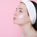 Side view of pensive female wearing headband while standing on pink background with cosmetic product on cheek and looking up
